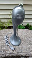 Silver vintage-style electric milkshake blender with metal mixing rod and power cord arrangement on a stone surface outdoors.
