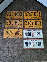 Overview showing six New York license plates, four yellow-orange vintage and two newer white with Statue of Liberty graphic, displaying various conditions.