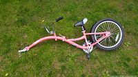 Pink Weeride Copilot bike trailer attachment lying on grass, showing curved frame, rear wheel, seat, and coupler hitch for attachment to an adult bicycle.