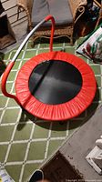 Top-down view of small round kids trampoline with black jump surface, red padded edge, and red/white handlebar in indoor setting.