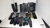 Lot of 27 assorted TV remote controls displayed on flat surface, showing a variety of models and conditions.