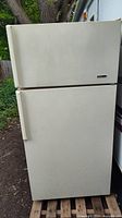 Front view of the used cream-colored Frigidaire Elite top-freezer refrigerator standing on a wooden pallet outdoors.
