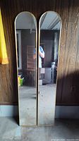 Two tall wall mirrors with gold-tone frames and rounded arch tops, leaning against a wood-paneled wall in a garage-like setting.