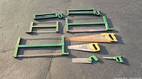 Photo showing nine vintage saws including four hand saws with wooden handles and five green metal frame saws laid out on concrete.