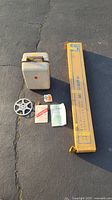 Lot displayed on pavement showing Kodak Showtime 8 projector in white carrying case, reels, manuals, SUPER 18 stand box, and screen box.