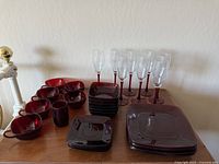 Full set of vintage 1960s Royal Ruby glassware including plates, bowls, cups, tumblers, and clear champagne flutes with purple stems arranged on a surface.