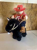 Front angle of Cabbage Patch Kid doll wearing cowboy outfit mounted on black plush horse with reins and saddle.