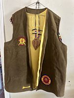 Brown vintage YMCA Indian Guides vest with yellow interior lining displayed hanging. Two colorful embroidered patches visible on the front: a star shape with 'Indian Guides' text and a circular patch with 'Indian Guides' text. Leather heart ornament with 'Full Moon' text hanging around the neck.