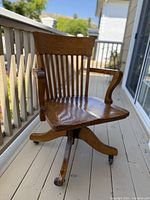 Front angle view showing slatted backrest, curved armrests, and contoured seat on oak swivel base with casters.
