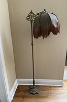 Full view of tall antique Victorian style floor lamp with ornate metal base and metal mesh lampshade with fabric fringe.