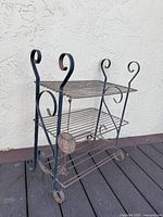 Three quarter view of black iron three-tier plant holder shelf with decorative scroll sides and owl ornament at front left.
