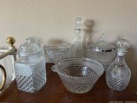 Wide view of the entire set of cut glass items showing ice bucket, decanters, large bowls, and storage containers arranged on wooden surface against a wall.