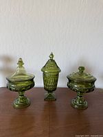 Full view of three green glass candy dishes with lids on a wooden surface against a white textured wall.