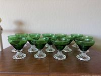 Front view of 14 green sherbet/champagne glasses arranged in rows on a wooden table, showcasing the deep green bowls and clear twisted stems.