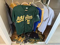 Green Oakland Athletics jersey with number 5, multiple hanging shirts, Nike sneakers, black A's beanie and pom-poms on floor