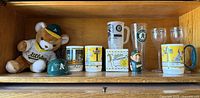 Vintage Oakland Athletics memorabilia collection displayed on wooden shelf including teddy bear, mini helmet, multiple mugs, clear drinking glasses, and bobblehead.
