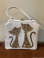 Front view of vintage white cat purse showing two decorative cats with jewel embellishments and floral accents on a glossy lucite-like surface, with white handle and gold-tone clasp.