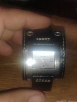 Front view of Fossil wristwatch showing square silver-tone face and black leather cuff band with metal rivets and white stitching, held by hand.