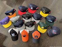 All 16 baseball hats laid out on dark wood floor from above showing various team logos and color combinations.