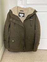 Front view of the olive green BCBG Generation women's jacket showing zippered chest pocket, two lower flap pockets, snap button closures, and sherpa fleece lining inside the hood and jacket.