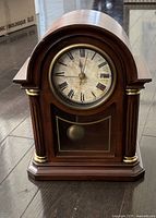 Front view of wooden mantel clock showing clock face with Roman numerals and brass accented wooden case with arch top and lower pendulum window.