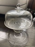 Full view of glass cake dish showing dome lid, round patterned plate, and pedestal stand on hardwood floor.