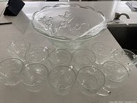 Clear glass punch bowl with floral embossing, 10 matching cups, and plastic spoon on countertop.