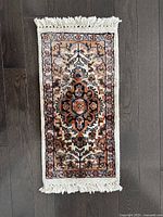 Mini Persian carpet laid flat on dark wooden floor showing full top view with fringed tassels on both ends and detailed central medallion pattern with complementary borders.