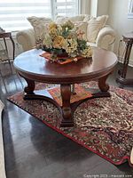 Photo showing a round dark wood coffee table with carved legs and stretcher base in a living room setting on a patterned area rug.
