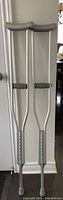 Pair of gray aluminum crutches standing side by side with padded underarm supports and hand grips, adjustable height with numbered settings visible, rubber tips at bottom