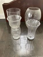 Four assorted clear glass vases set against a dark floor and wood furniture: one tall cylinder, one short rounded, one flared with vertical ridges, one tall rounded vase.