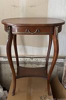 Full front view of wooden oval side table showing drawer with brass handle, curved legs with brass accents, and lower shelf