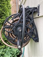 Black ornate water hose holder with light brown hose coiled on it, mounted on white siding next to greenery.