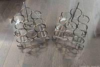Two metal wine holders placed side by side on a dark wood floor, showing the top and front view with circular slots for bottles and central spiral handle.