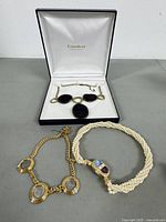Three necklaces laid out on a grey surface, one in an open white box branded Gundert showing gold-tone and black elements, one multi-strand white beaded necklace with a decorative clasp, and one gold-tone necklace with textured gold settings.