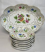 Set of 6 nut bowls stacked with large serving bowl behind, showing matching floral and gold hand painted designs with scalloped edges.