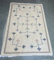 Top-down photo of the entire handwoven Kilim area rug showing the geometric floral pattern with crosses and arrow-like motifs in muted colors on a beige background with fringed edges.