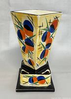 Front and angled view of the ceramic vase showing the Art Deco floral pattern with blue, orange, green, and black colors on a yellow checked background. The base is square and black glazed with similar decorative motifs.