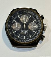 Front view of watch showing black dial with Heuer logo, three subdials, date window at 3 o'clock, tachymeter scale and scratched crystal