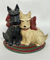 Front and side view of the cast iron doorstop depicting two dogs with a red plaid bow sitting on a red and green striped oval base.