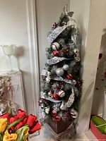 Full view of the decorated Christmas tree showing red and silver ornaments and white ribbon on green and silver-tinged branches, standing on a deep red plastic base.