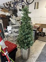 Artificial Christmas tree standing in a garage. The tree is tall and full with green plastic pine needles. It is growing out of a tall, gold/bronze colored plastic pot.