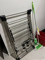 Folded aluminum drying rack leaning against wall with mop and cleaning supplies nearby.