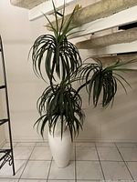 Front view of artificial plant with three main branches of long green plastic leaves in tall white porcelain vase, placed on tiled floor near stairs.