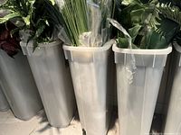 Multiple bunches of artificial plants in tall white plastic containers with varied green and white colors visible, including grass, leafy plants, and flowers.