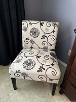 Front view of the accent chair showing the floral and swirl pattern upholstery in cream and black tones, black wooden legs, and slight fabric mark visible.