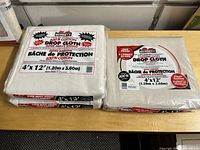 Three drop cloths on a wooden table. Two Bennett cotton drop cloths stacked and one Tremor laminated canvas drop cloth.