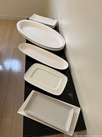 Eight white ceramic serving dishes arranged on black surface, various oval and rectangular shapes, glossy finish