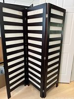 Three-panel black wooden frame room divider with beige fabric inserts shown standing in folding position in living space.