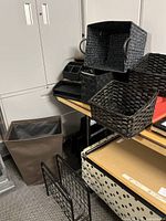 Multiple black wicker and woven baskets displayed on and under a wooden table with metal legs. Includes woven and wicker materials, some with metal handles, a large brown fabric bin, and a red decorative box.